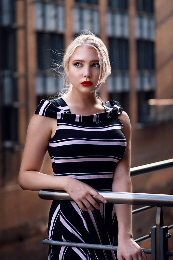 Young woman with blonde hair and red lipstick wearing a striped dress, leaning on a railing.